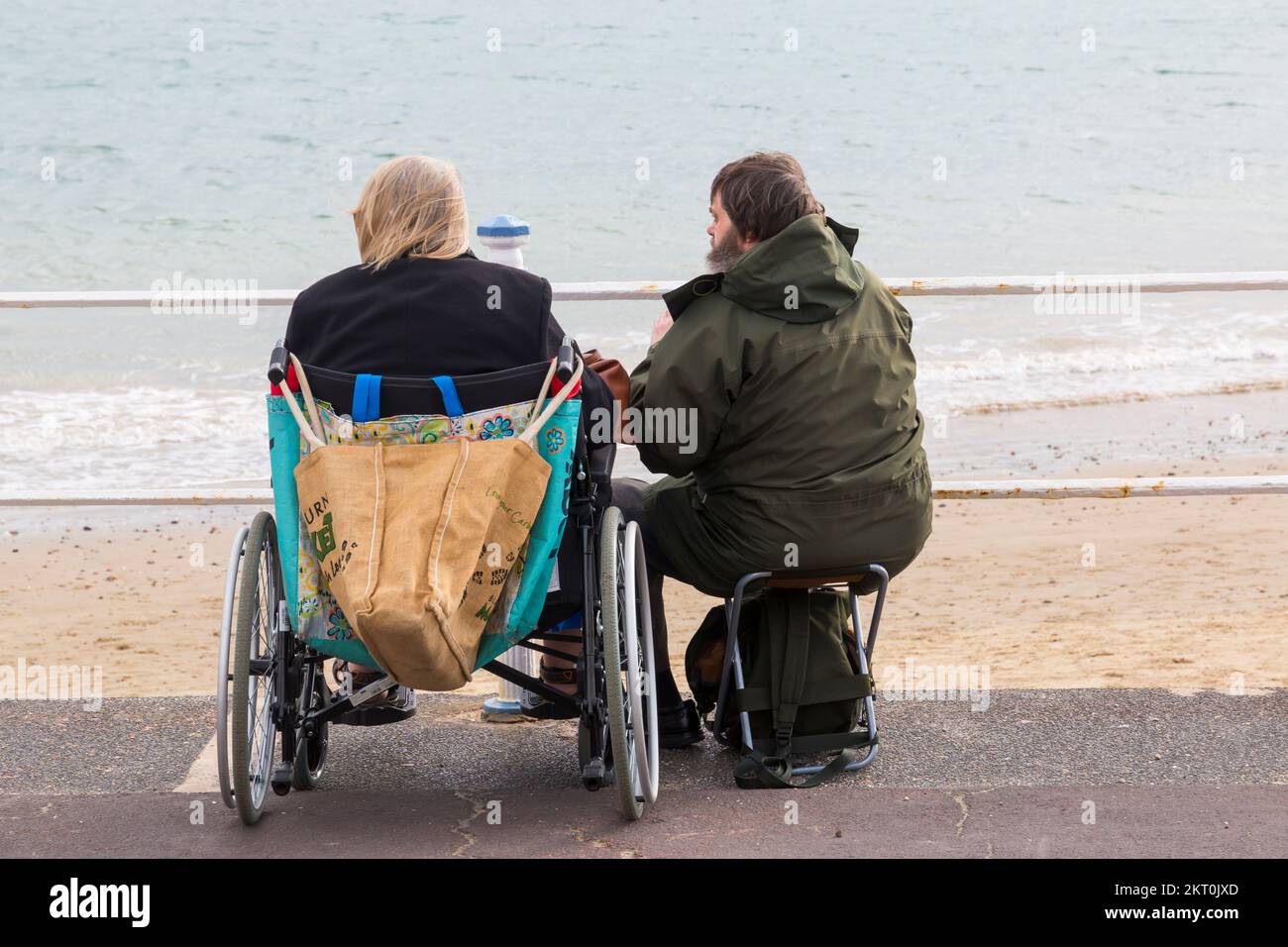 Couple sitting on promenade esplanade, woman in wheelchair, man in fold up seat, at Weymouth, Dorset UK in October Stock Photo
