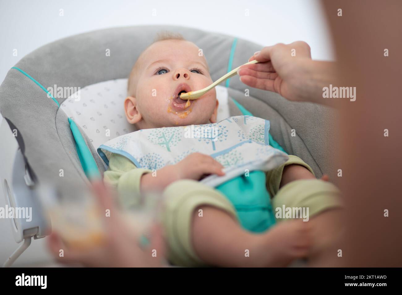 Mother spoon feeding her baby boy child in baby chair with fruit puree. Baby solid food introduction concept Stock Photo