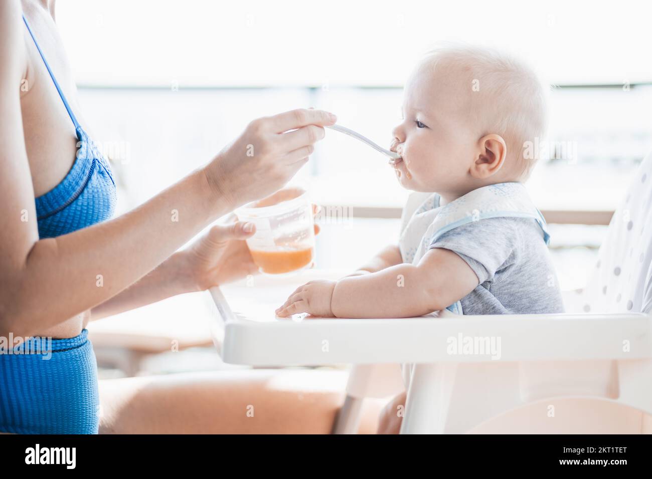 Mother spoon feeding her baby boy child in baby chair with fruit puree on a porch on summer vacations. Baby solid food introduction concept Stock Photo