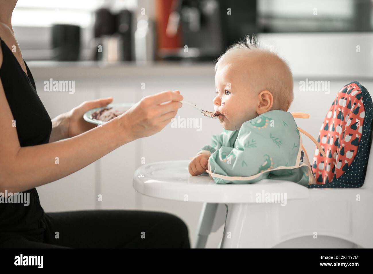 Mother spoon feeding her baby boy child in baby chair with fruit puree in kitchen at home. Baby solid food introduction concept Stock Photo