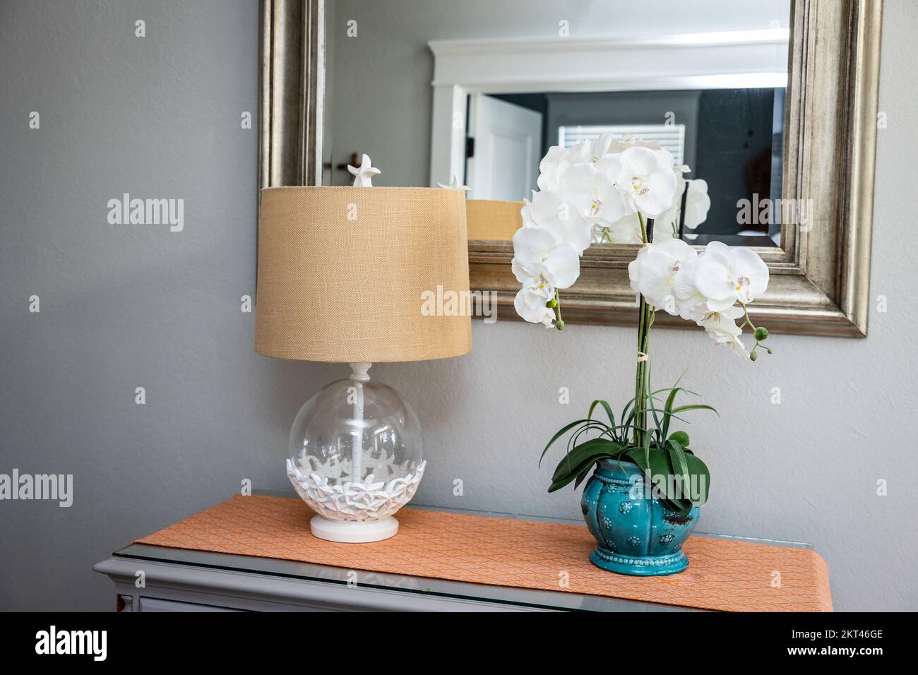 Front entryway table decor of a lamp and orchid with a mirror and orange runner on a gray dresser table Stock Photo