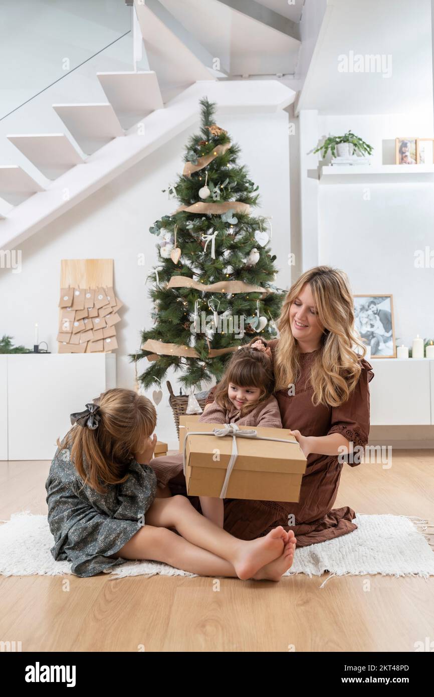 Little sisters with her mother opening a gift near the Christmas tree at home Stock Photo