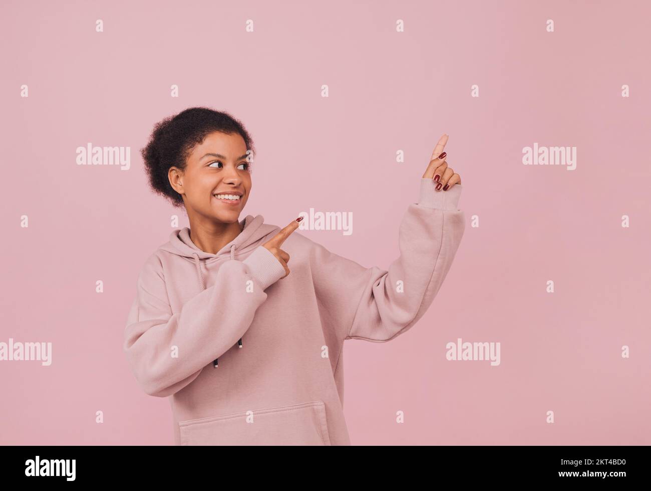 Smiling girl with pointing gesture. Advertising concept. Portrait of happy black woman in pink hoodie against pink background Stock Photo