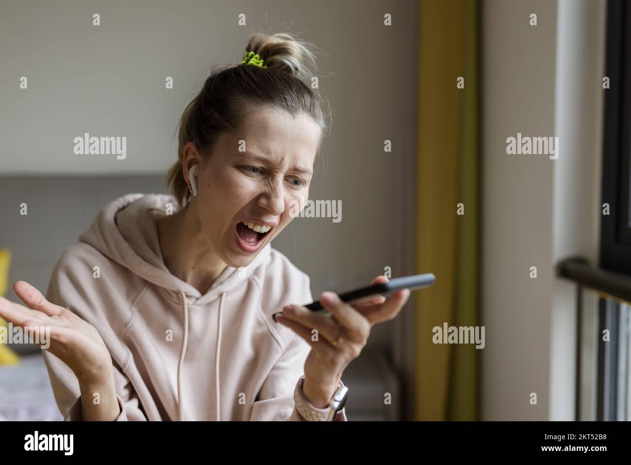 Young caucasian woman using mobile phone and wireless headphones, resting, sitting on bed at home and listening podcast or clubhouse - voice-only Stock Photo