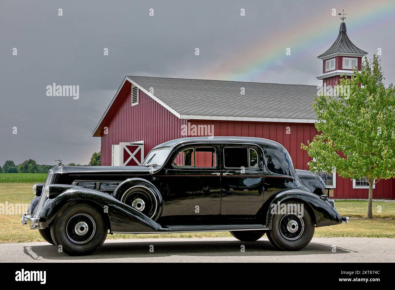 Vintage black automobile by a red farm barn with rainbow Stock Photo