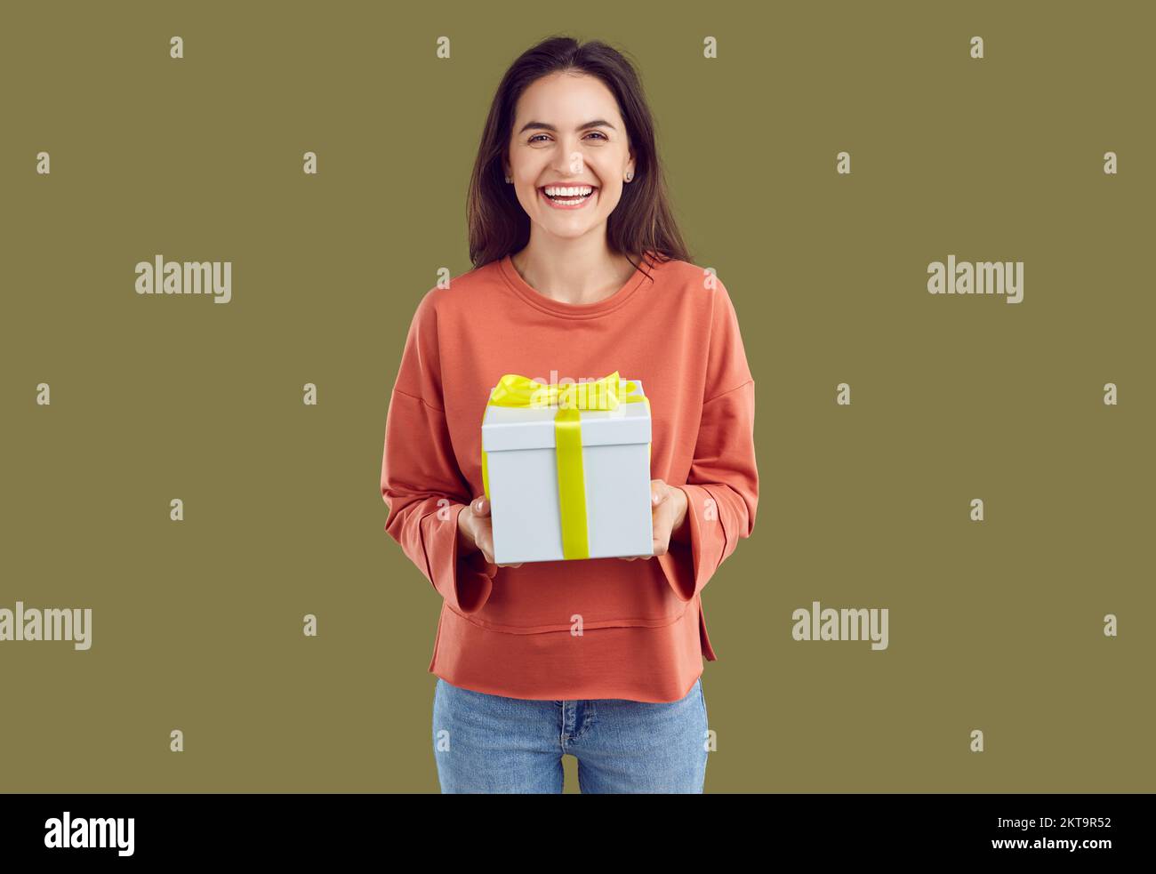 Happy laughing brunette woman in red sweatshirt is holding a present in hands on khaki background. Stock Photo