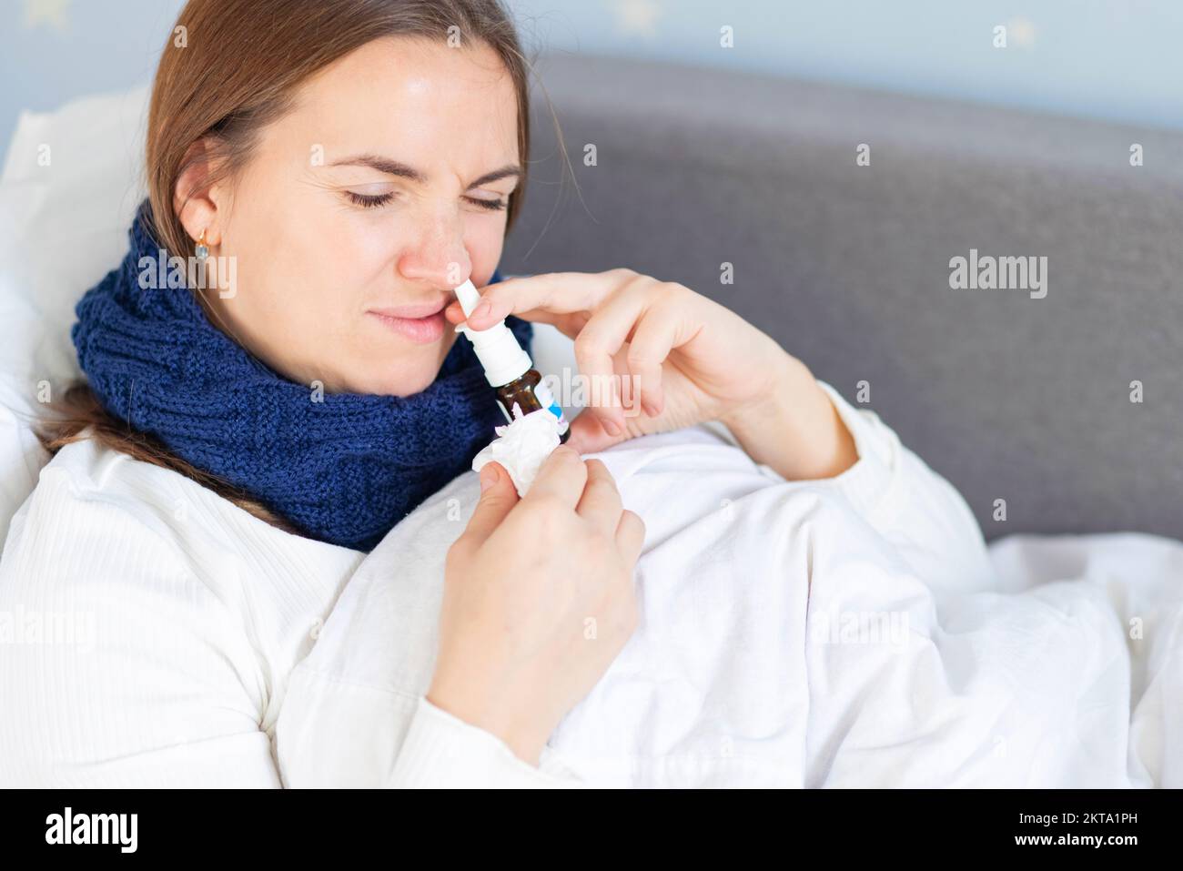 Young woman with bad cold lying in bed under blanket and using nasal