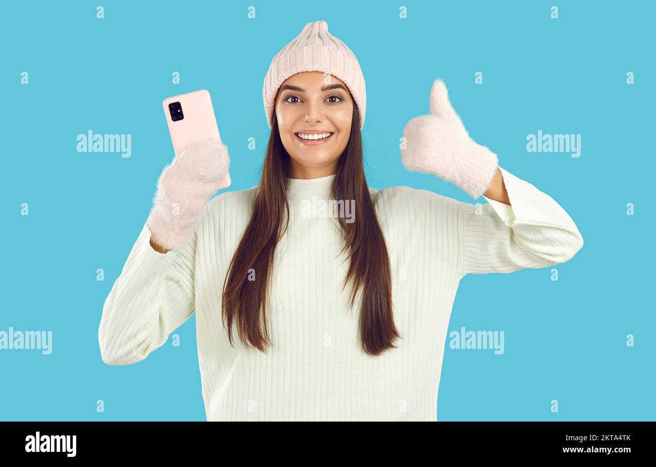 Happy woman in winter hat and mittens advertising modern cellphone and giving thumbs up Stock Photo
