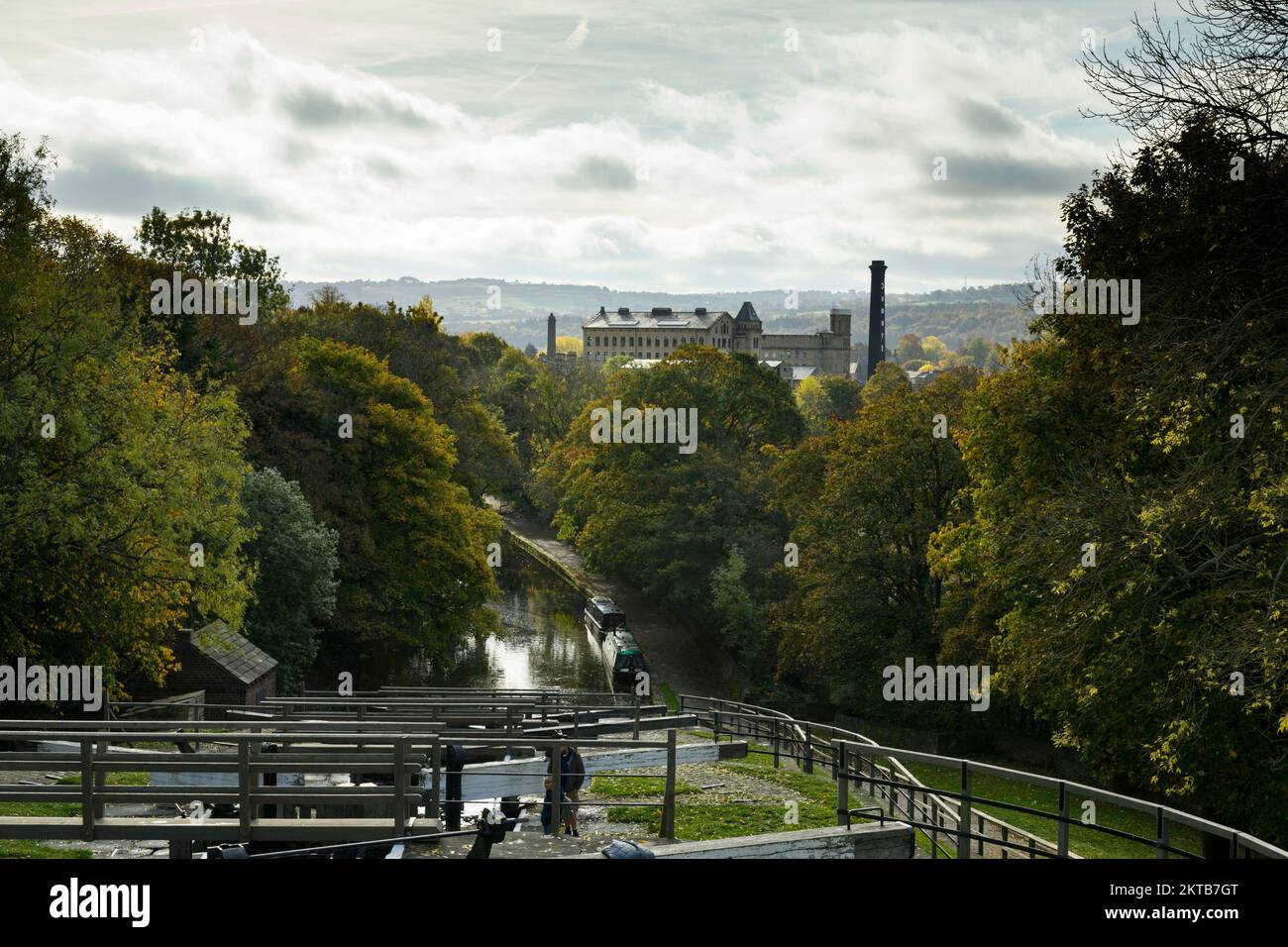 2 people by lock gates, boats moored, sunlit Damart mill, autumn colour - Bingley Five Rise Locks, Leeds & Liverpool Canal, West Yorkshire England UK. Stock Photo