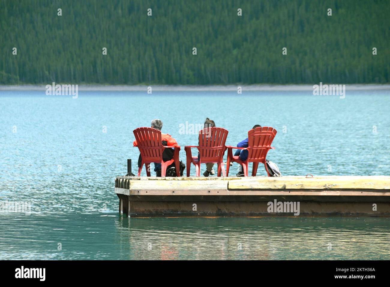 Banff, Alberta, Canada - June 2018: Three people sitting on chairs on a wooden jetty floating on the waters of Lake Minnewanka Stock Photo