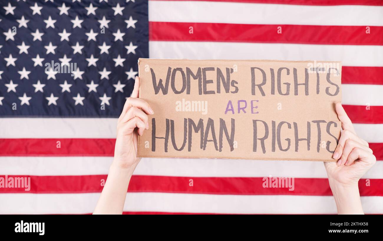 Young woman protester holds cardboard with Womens Rights Are Human Rights sign against USA flag on background. Feminist power. Equal opportunity Women Stock Photo