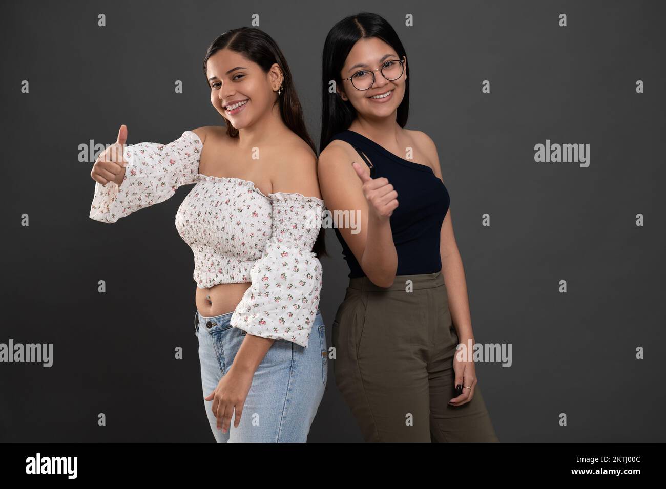 Two young girls with thumb up  smiling on grey studio background Stock Photo