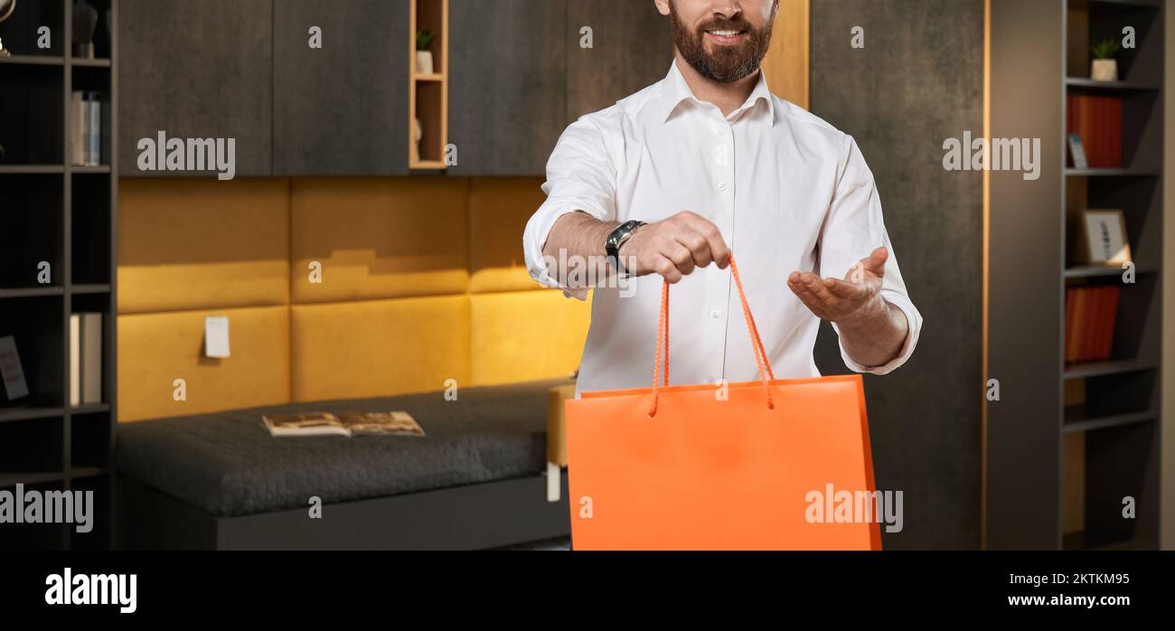 Unrecognizable male retailer in shirt giving orange shopping bag to camera in furniture store. Crop view of smiling bearded seller, offering purchase, Stock Photo