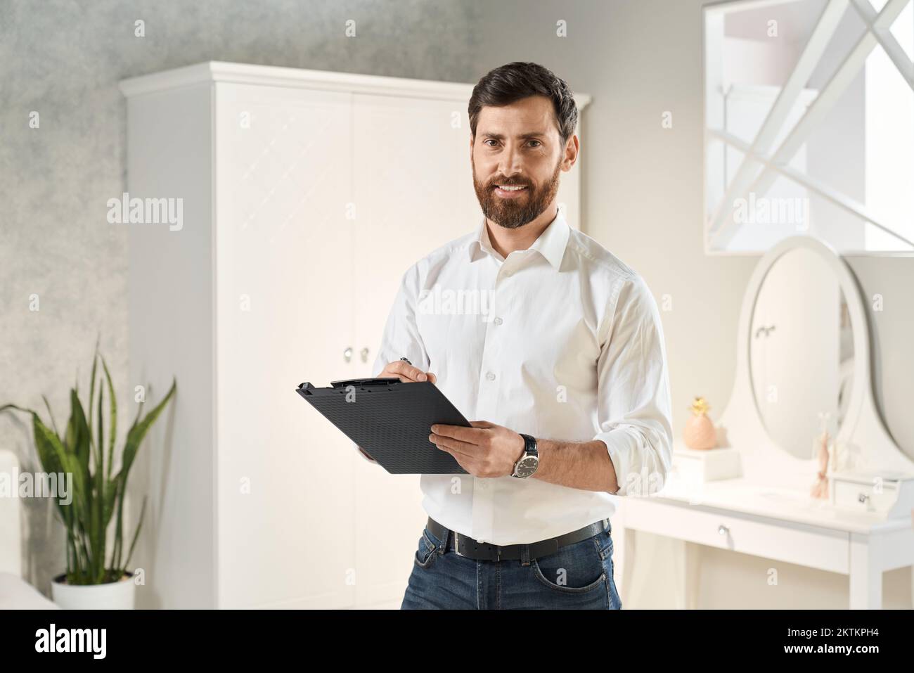 Professional retailer standing with clip-on tablet, while smiling at camera in exhibition center. Portrait of elegant male expert working, with bedroo Stock Photo