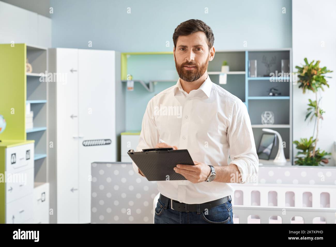 Handsome salesman with black tablet looking at camera, while standing in showroom. Portrait of attractive male retailer in shirt making notes, while w Stock Photo