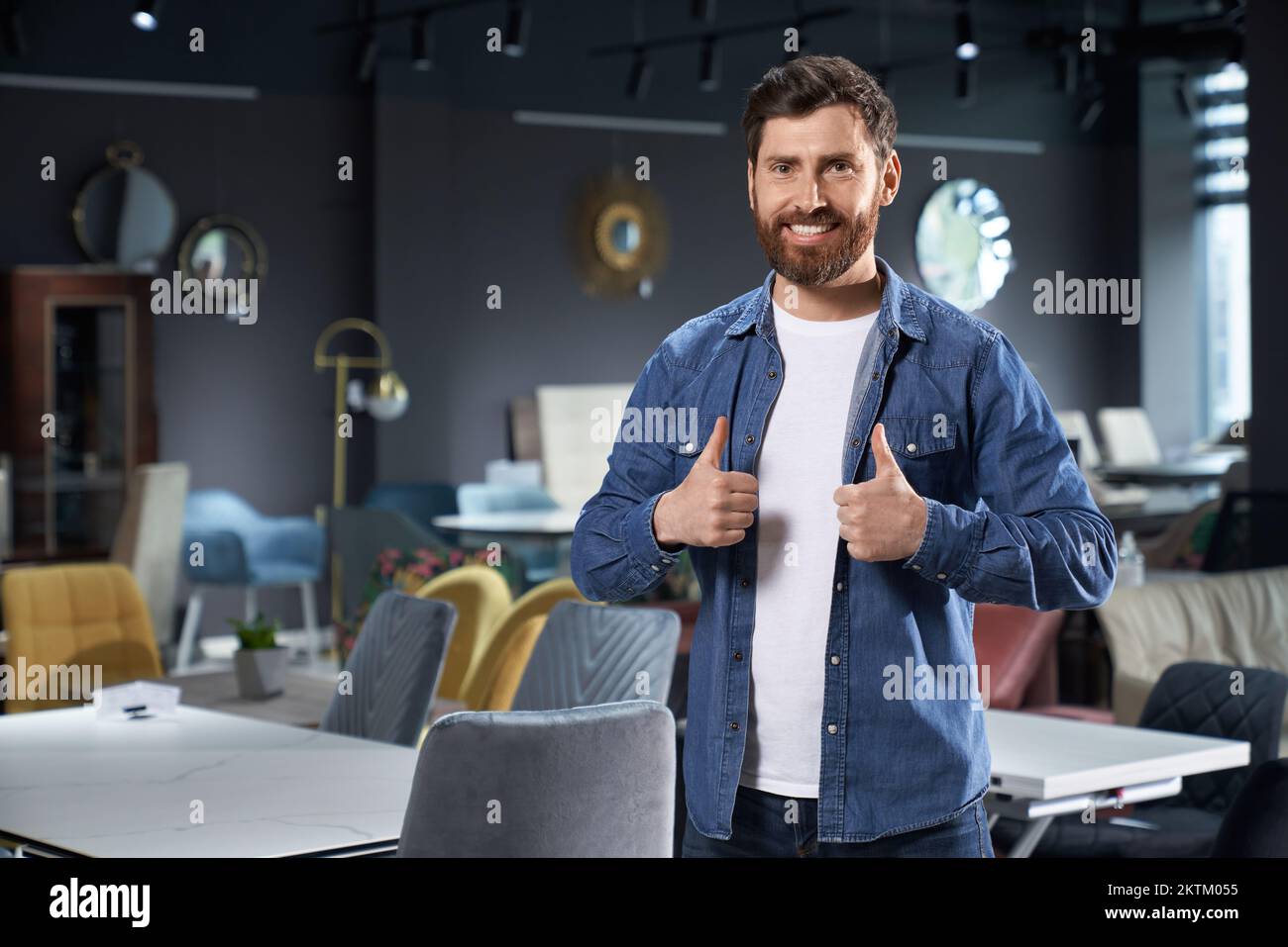 Handsome bearded man showing thumb up, recommending, while standing in furniture store. Portrait of cheerful elegant guy making like gesture, while lo Stock Photo