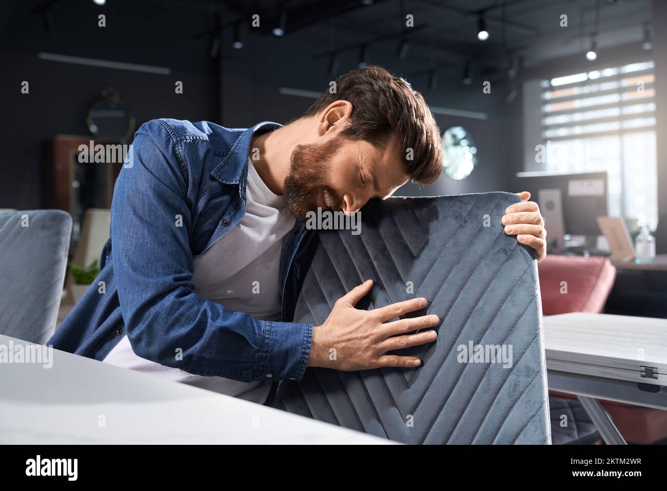 Romantic bearded man in casual outfit touching soft pattern, while leaning to chair indoors. Smiling brunet male with closed eyes hugging grey chair back, while sitting at table. Concept of emotions. Stock Photo