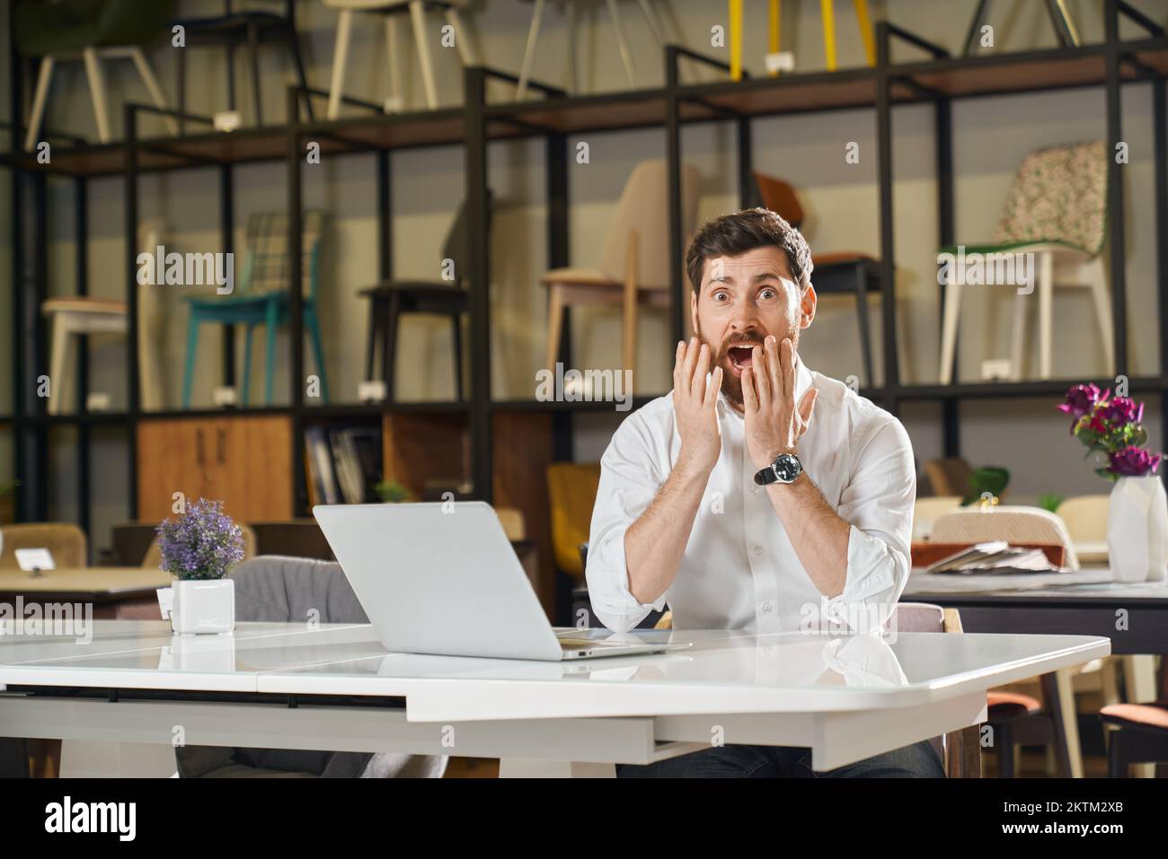 Amazed manager in white shirt browsing on laptop in furniture store. Portrait of shocked male retailer find out of profit results or career opportunities, while working indoors. Concept of emotions. Stock Photo