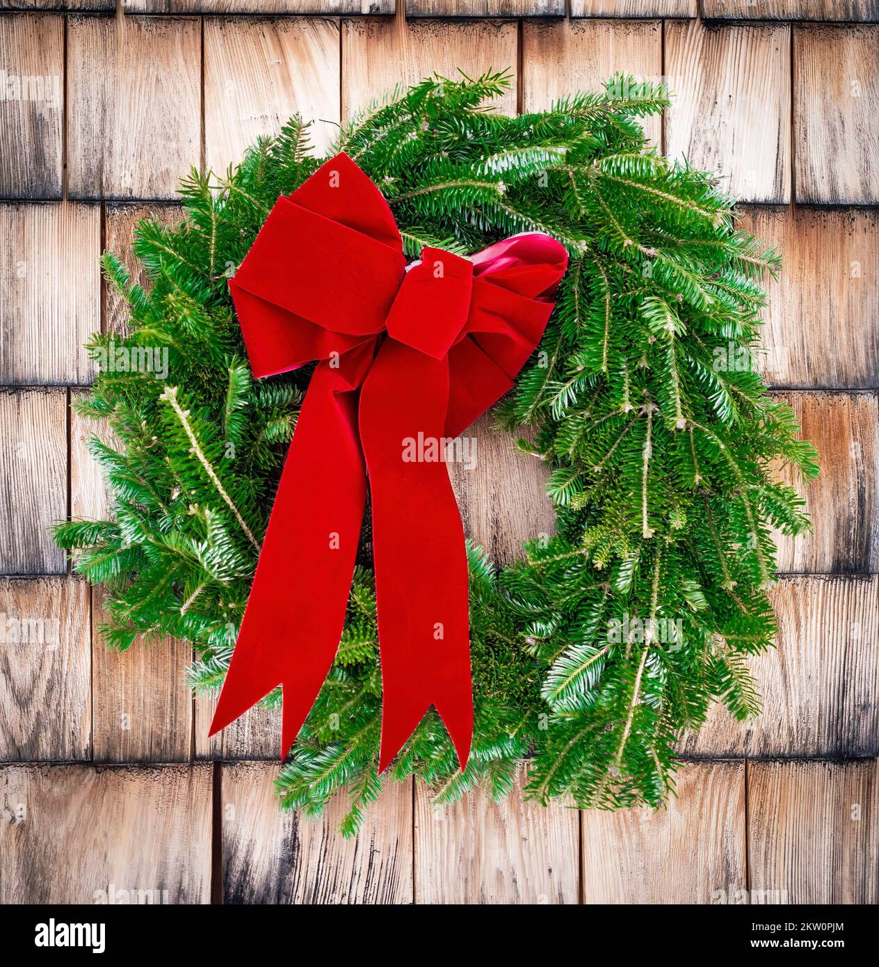 Pine needle Christmas wreath with big red bow on weathered cedar shake shingled house. Hint of snow. Rustic, natural, traditional country decoration. Stock Photo