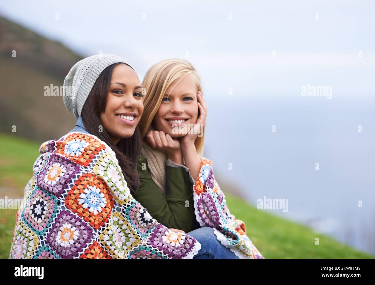 Friendship keeps us warm. Two young female friends sitting wrapped up in a blanket outside. Stock Photo