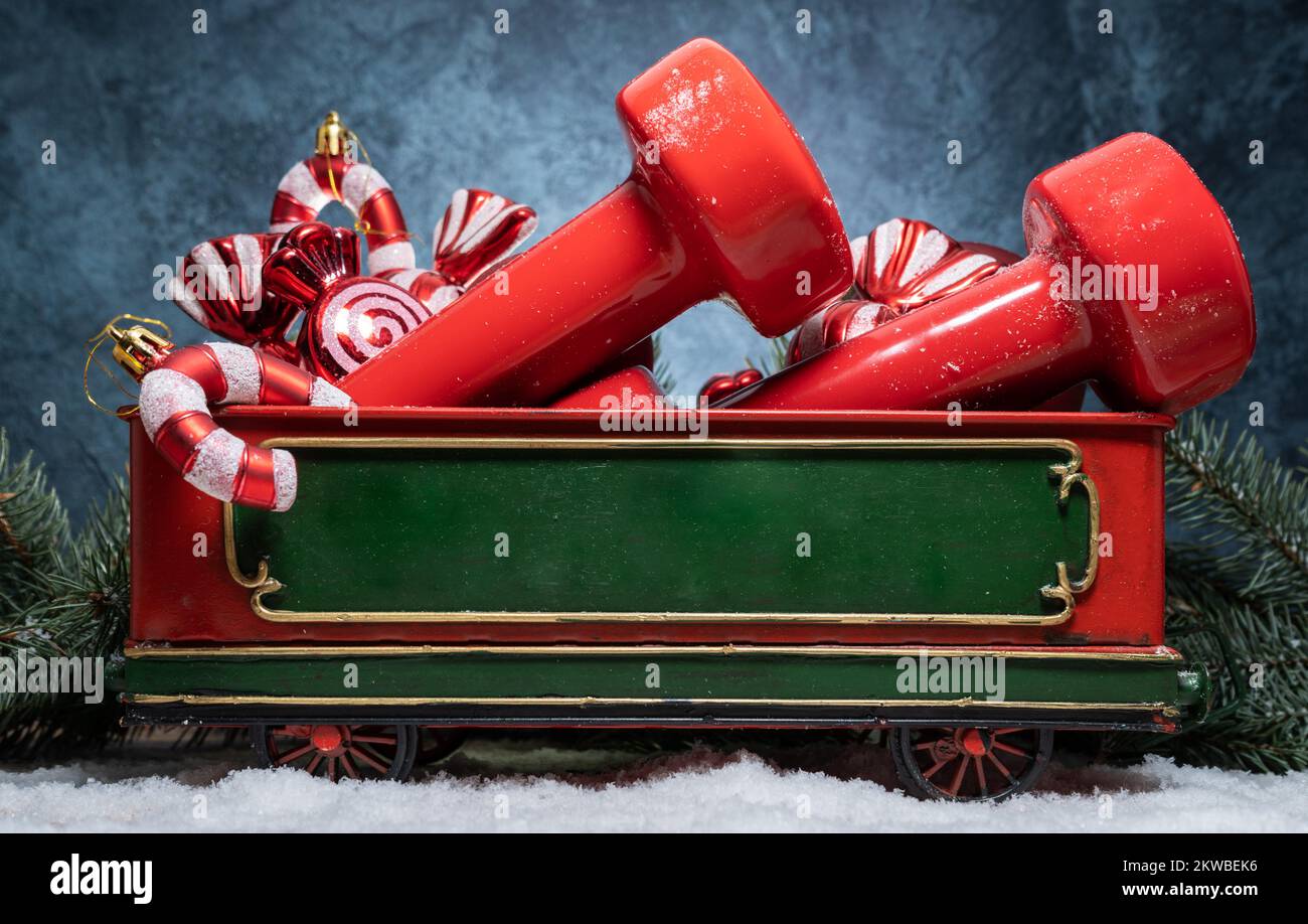 Gym dumbbells, candy canes decorations, ornaments in Christmas train car wagon carriage. Fitness holiday season winter composition with copy space. Stock Photo
