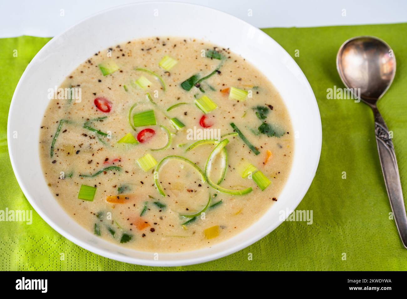 Dense leek soup with chilli pepper in white plate, spoon, fresh green background, closeup. Stock Photo