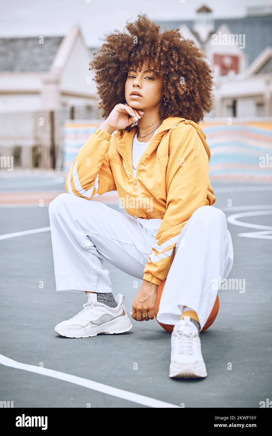 Trendy, stylish and fashionable female basketball player sitting on a ball at the court against an urban background. Young, edgy and serious woman Stock Photo