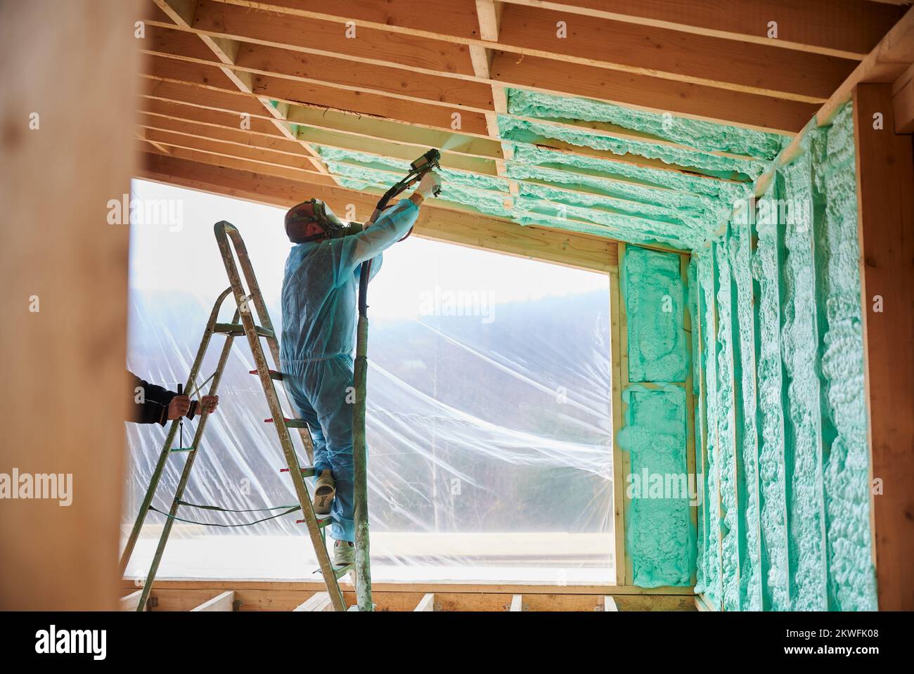 Male builder insulating wooden frame house. Man worker spraying polyurethane foam inside of future cottage, standing on ladder, using plural component gun. Construction and insulation concept. Stock Photo