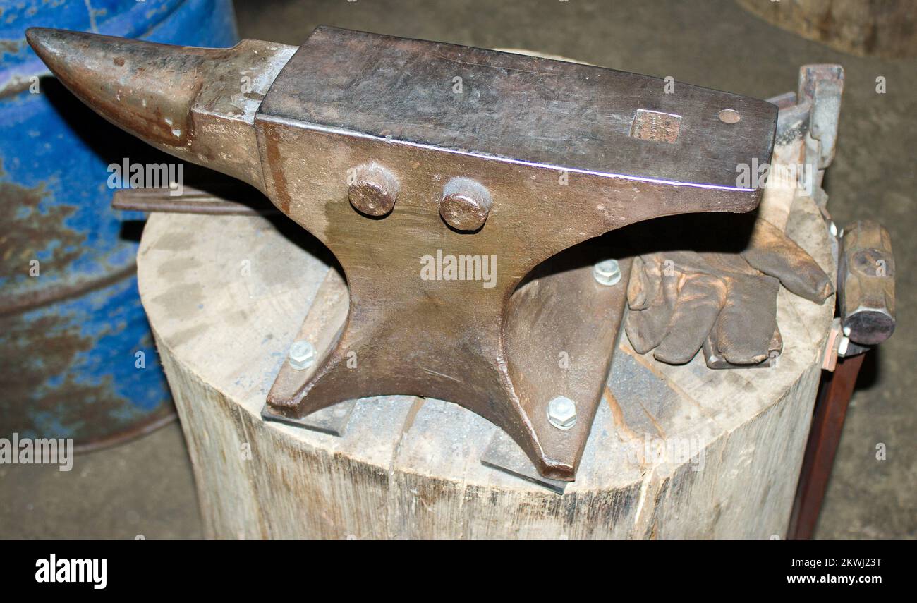 Old anvil, mounted on block of wood, used by blacksmith or forger in his workshop with hammer and glove Stock Photo