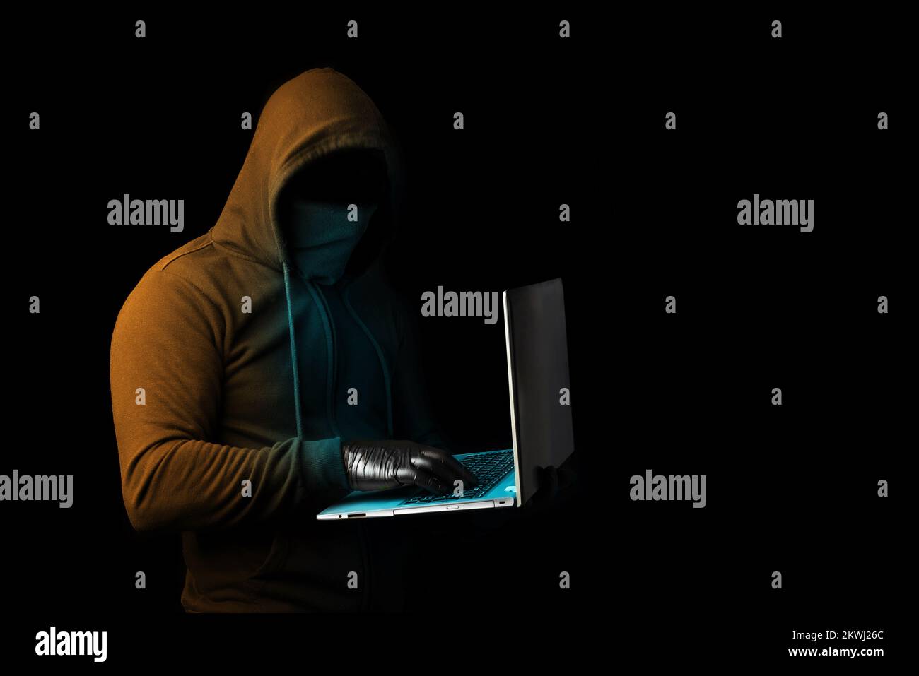 A hooded cyber criminal holding a laptop and scamming online. Cybercrime and phishing alert on the net Stock Photo