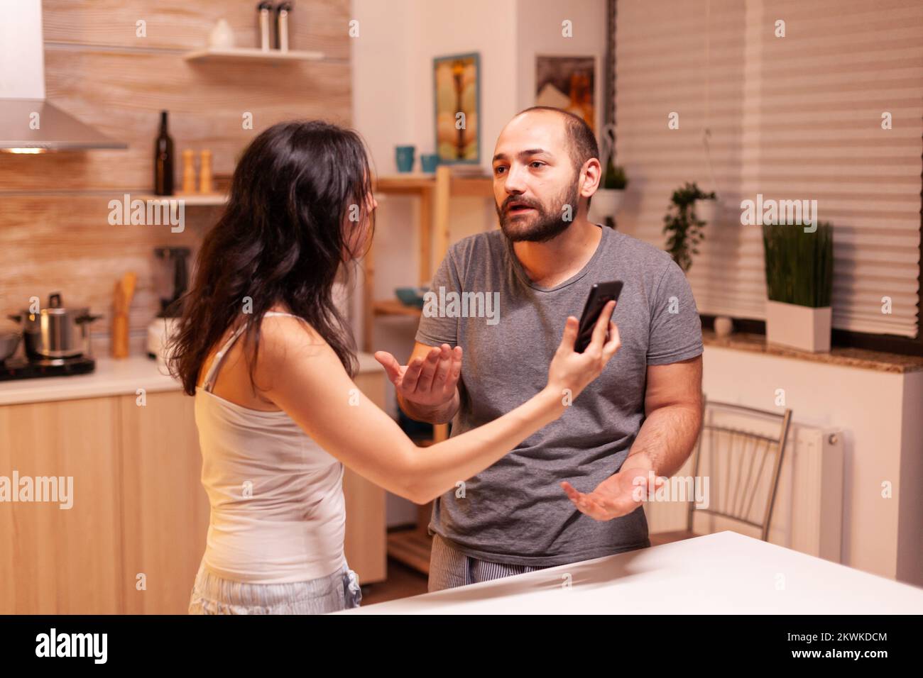 Husband caught cheating having a conflict with wife in home kitchen. Heated angry frustrated offended irritated accusing her man of infidelity showing him messages on smartphone yelling desperate. Stock Photo