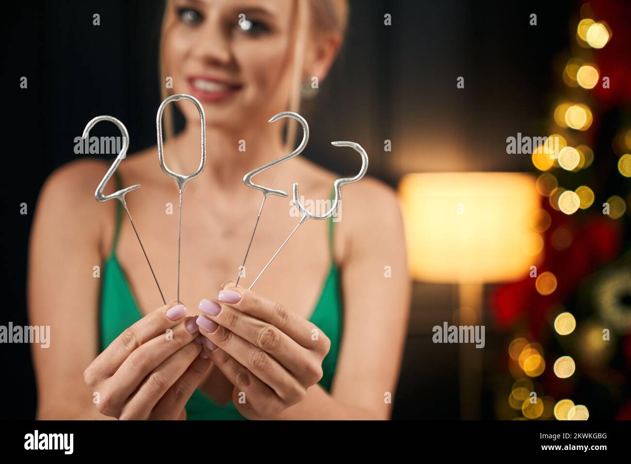 Front view of happy, cheerful lady with makeup and manicure, holding 2023 sparkles, showing. Sexy, young lady wearing green dress, smiling, Concept of New Year and celebration. Stock Photo
