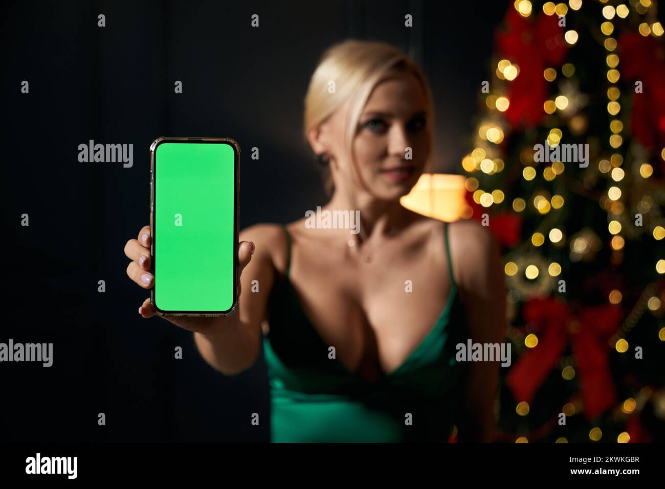 Front view of pretty, blonde woman holding smartphone, showing, advertising. Attractive, young lady wearing green dress, smiling. Concept of modern lifestyle and holidays. Stock Photo