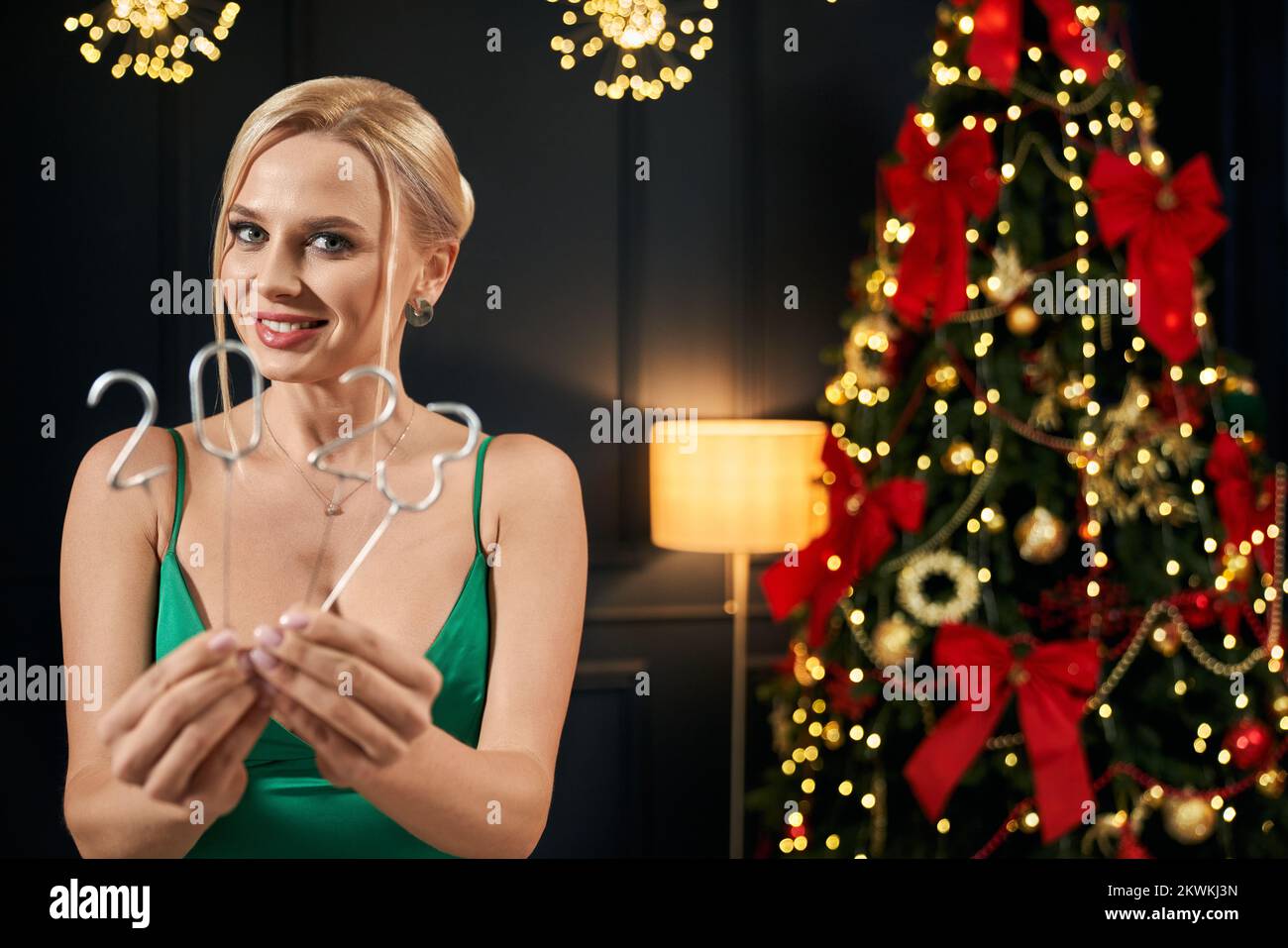 Front view of beautiful, blonde woman celebrating 2023 in decorated room. Sexy, young female wearing green dress, looking at camera, smiling. Concept of New Year and Christmas. Stock Photo