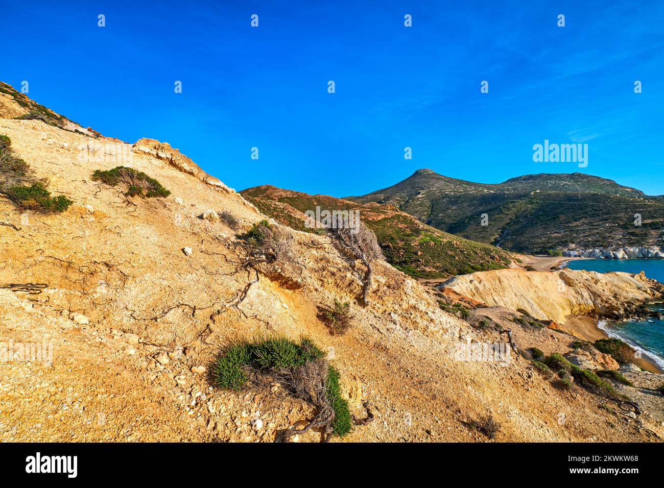 Natural rock formations of yellow cliffs on low sunlight, Milos, Greece. Weather-shaped slopes of rocks on Agios Ioannis beach at clear blue sky. Stock Photo