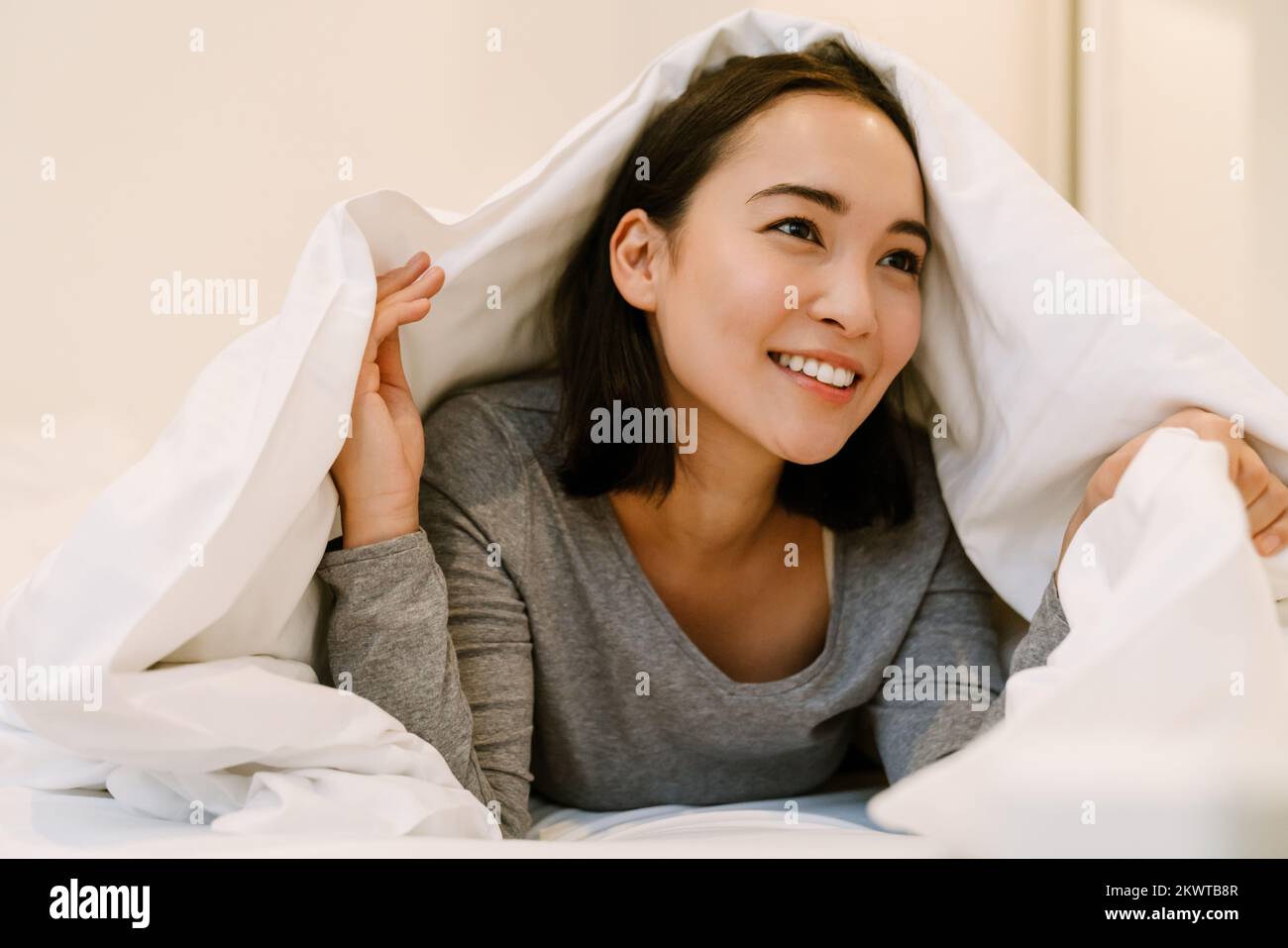 Young smiling asian girl in home suit lying on bed under covers at home