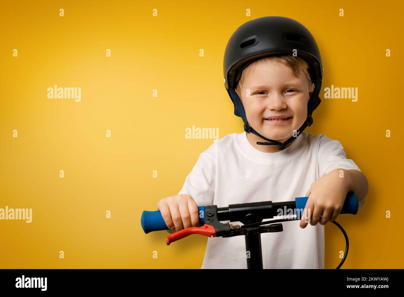 young smiling boy with helmet standing on kick scooter on yellow background with copy space Stock Photo