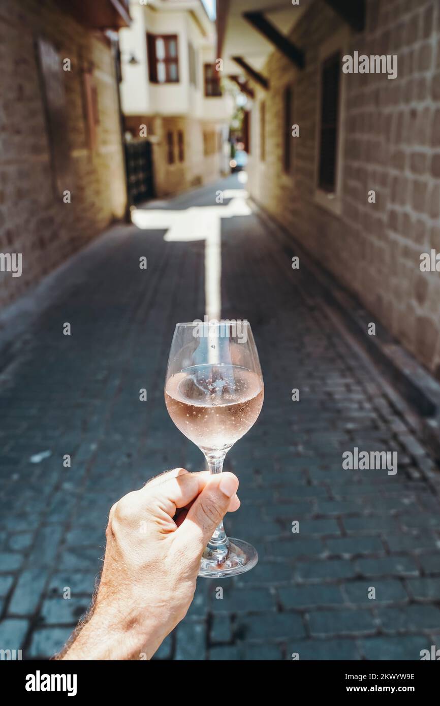 Glass of wine in hand. A glass of young fresh rose wine against the backdrop of a summer cafe in a Mediterranean seaside tourist town in the summer under sunlight. Summer, travel, lifestyle, relaxation, and enjoyment concept. High quality photo Stock Photo