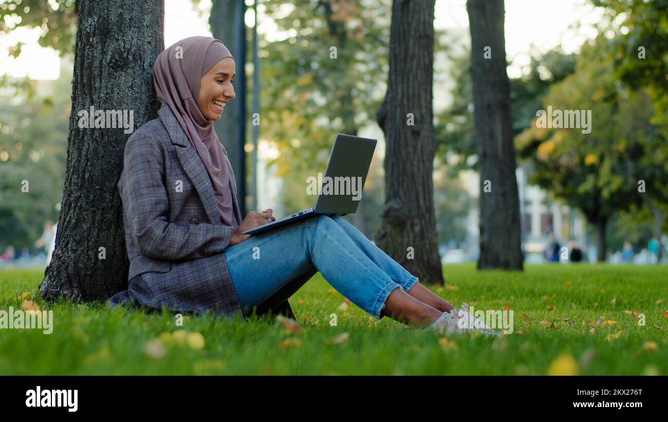 Happy muslim islamic girl business woman in hijab sitting near tree on green grass in city park chatting with friends family remote conversation Stock Photo