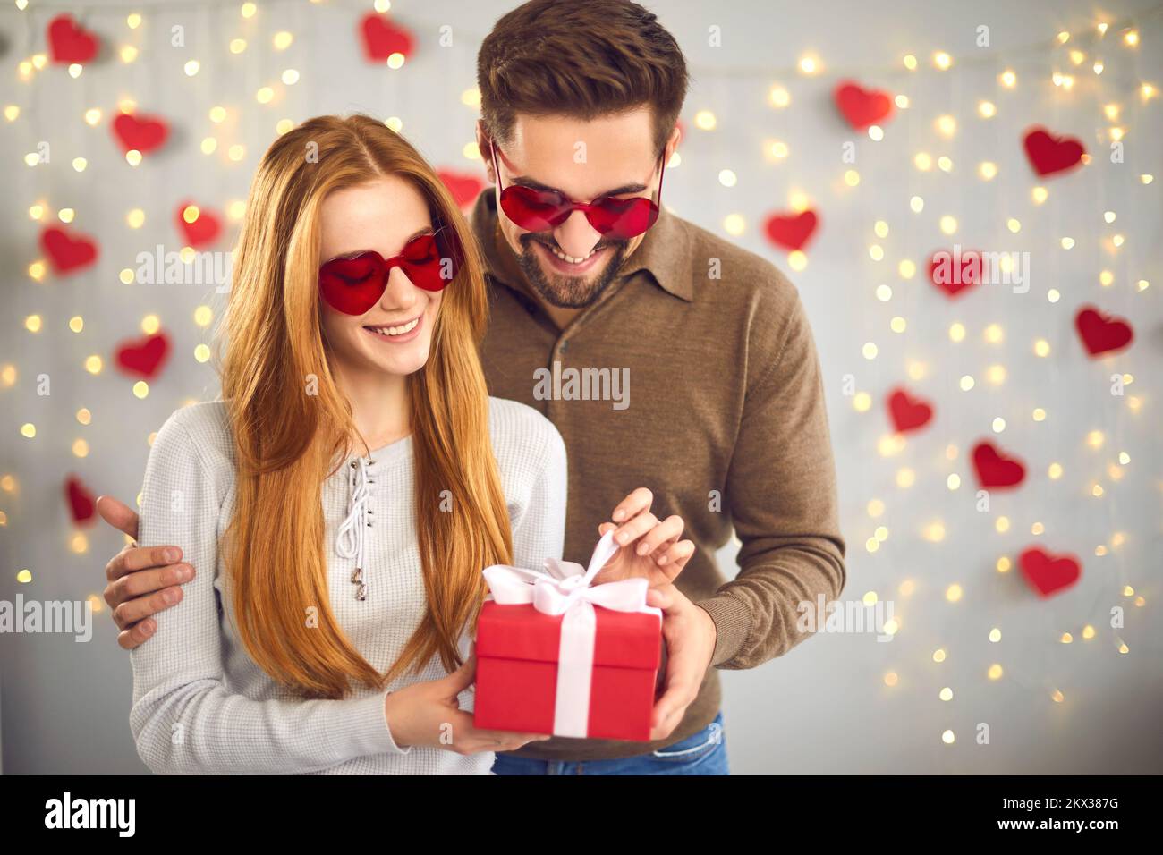 Loving couple unpacks a gift for Valentine's Day standing on a background of garlands and hearts. Stock Photo