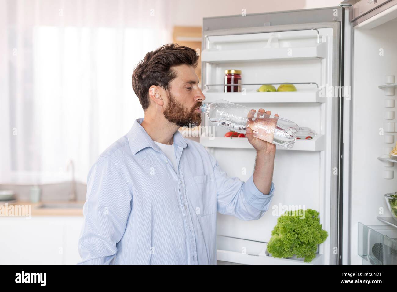 Adult thirst caucasian man with beard in white shirt opens refrigerator door, drinks water from bottle Stock Photo