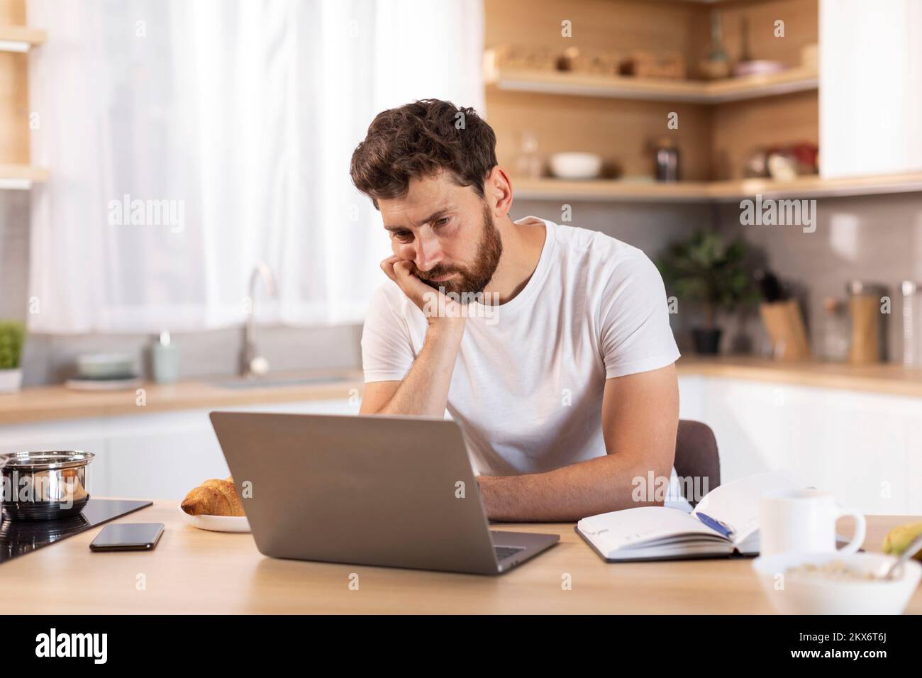 Boring tired sad adult caucasian man with beard in white t-shirt watch online lesson at laptop in modern kitchen Stock Photo