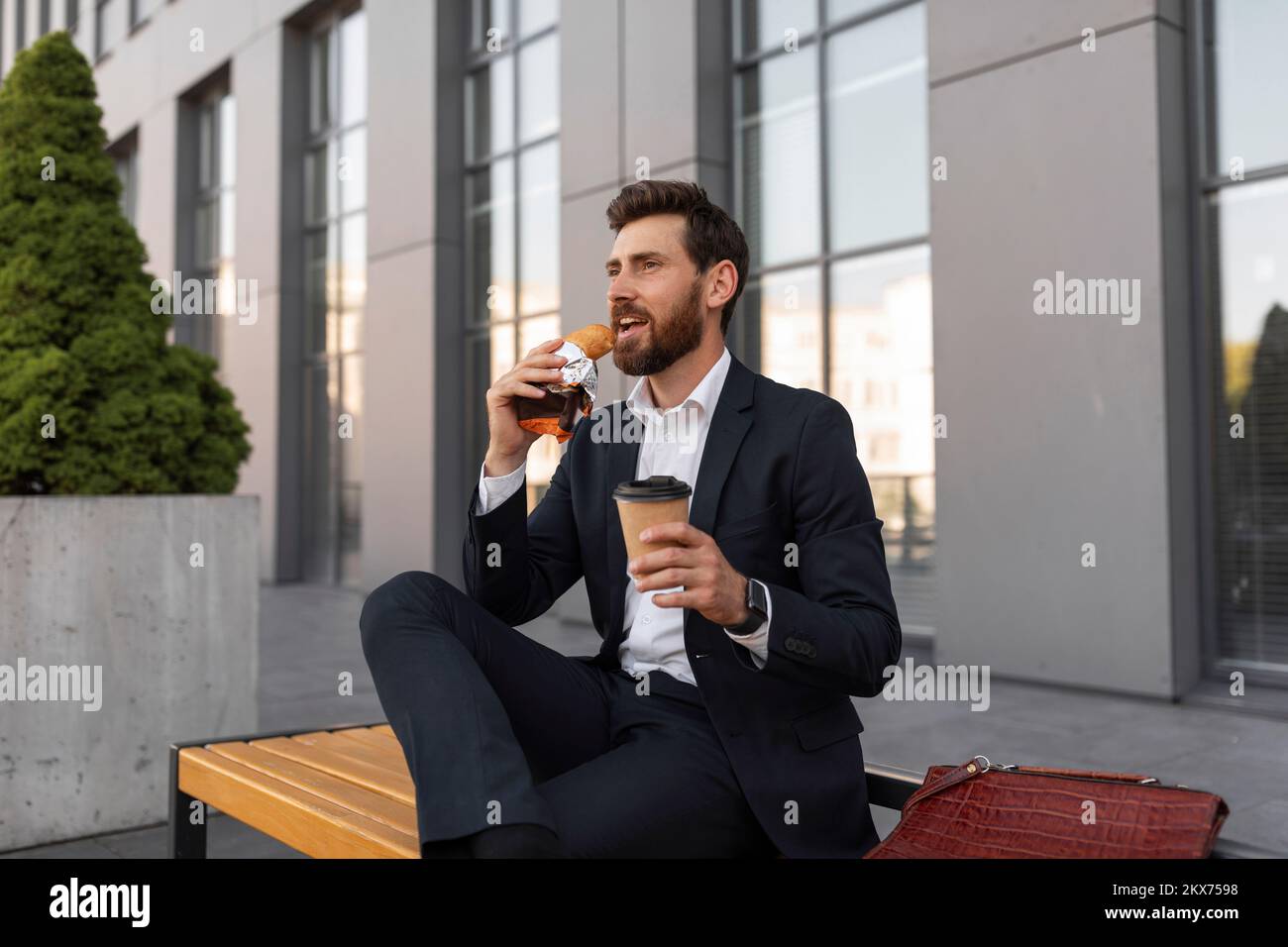 Happy hangry young european male manager with beard in suit with cup of coffee takeaway eats croissant Stock Photo