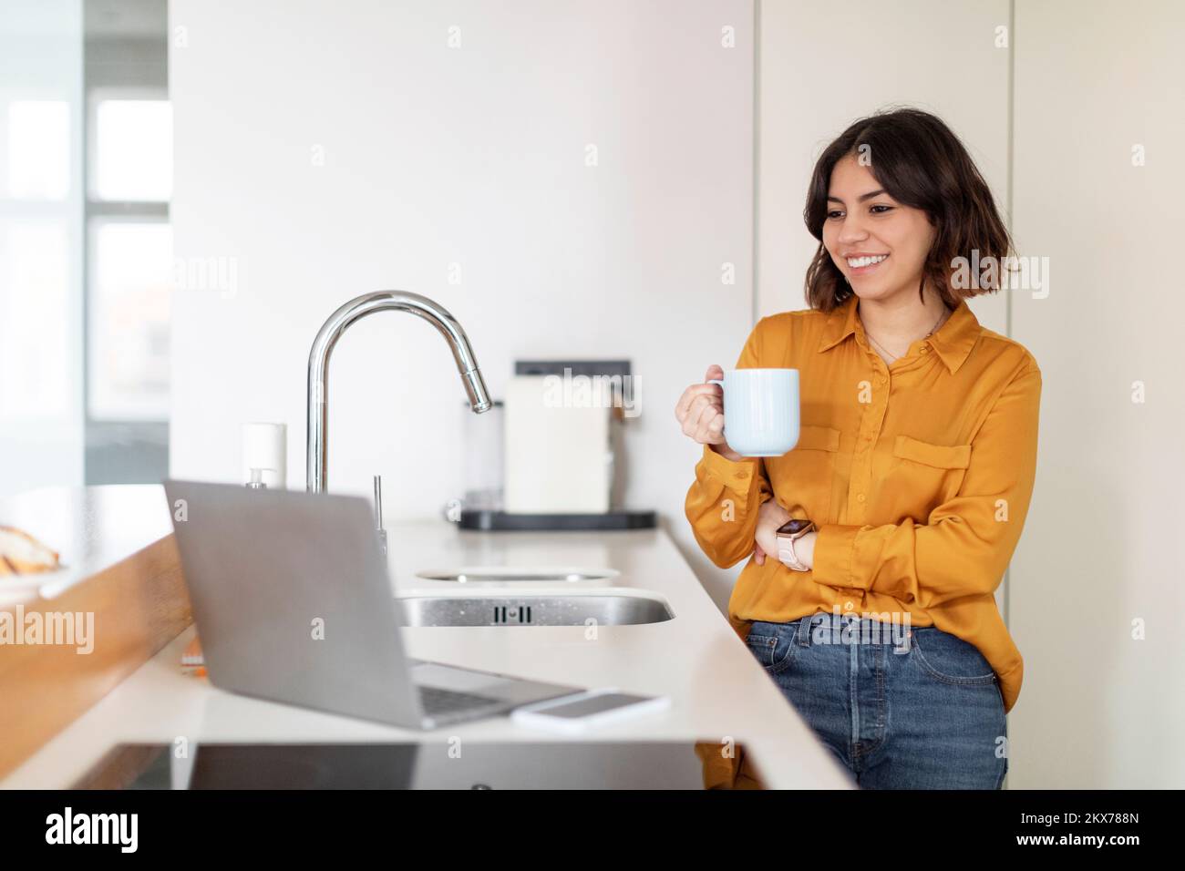 Young Smiling Arab Woman Using Laptop And Drinking Coffee In Kitchen Stock Photo