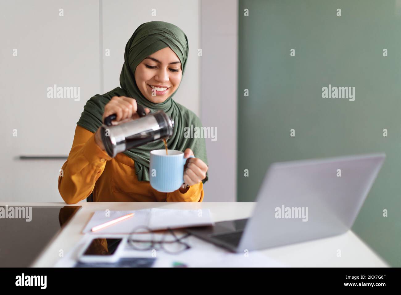 Smiling Muslim Female Freelancer Sitting At Desk With Laptop And Pouring Coffee Stock Photo