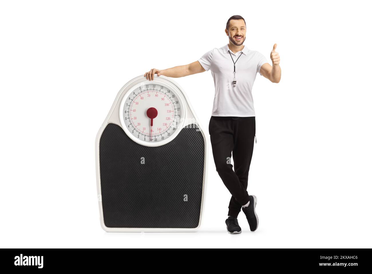 Fitness coach standing next to a weigh scale and showing thumbs up isolated on white background Stock Photo