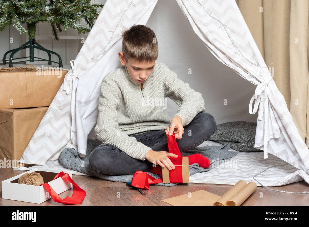 Gift wrapping. A teen child wraps Christmas gifts surprises in kraft paper with a rope, red ribbons. A serious boy is packing gifts for the family. Fa Stock Photo
