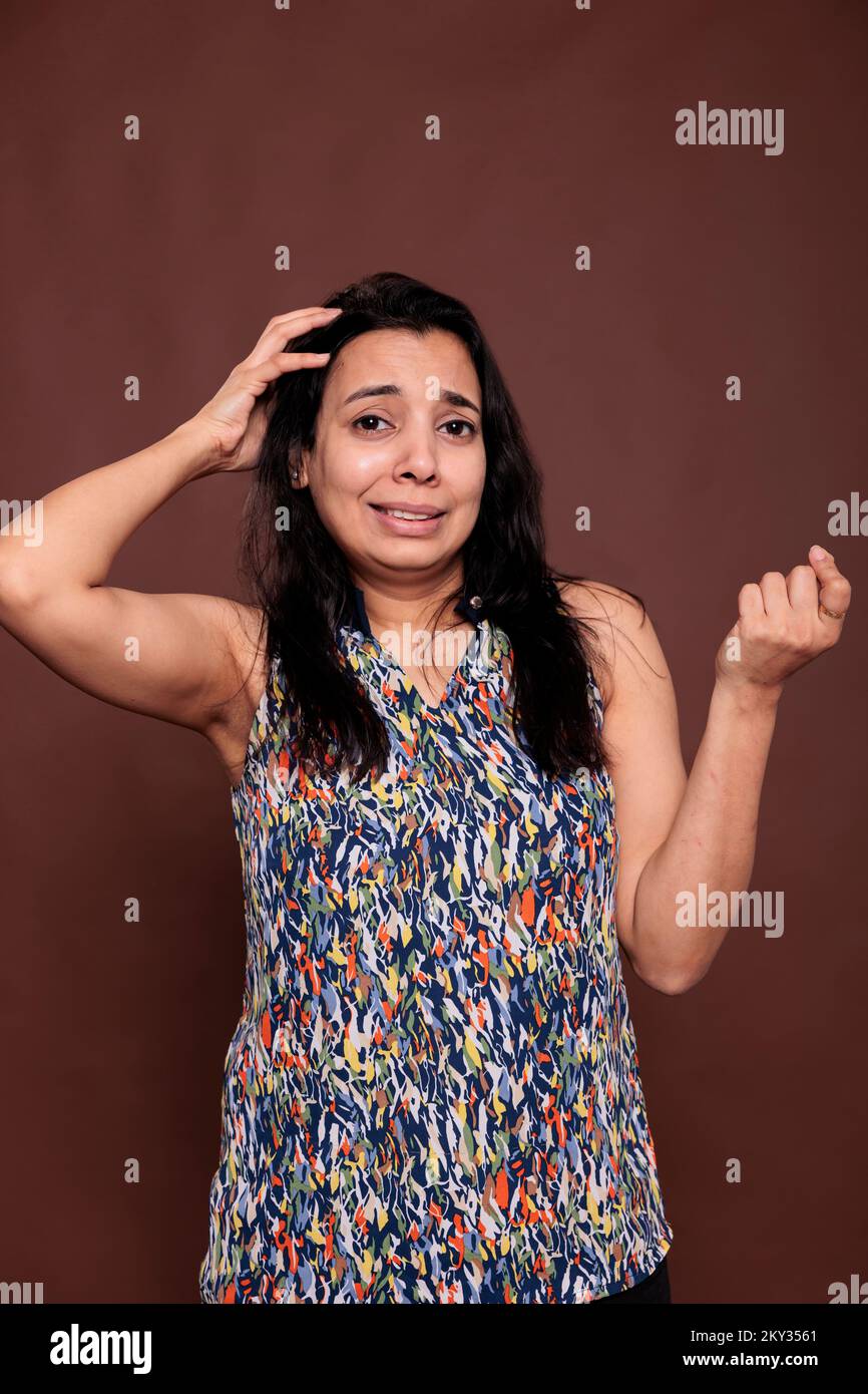 Thoughtful smiling indian woman rubbing head with confused facial expression portrait, awkward pose. Puzzled lady doubting, uncertain person thinking, looking at camera, studio medium shot Stock Photo