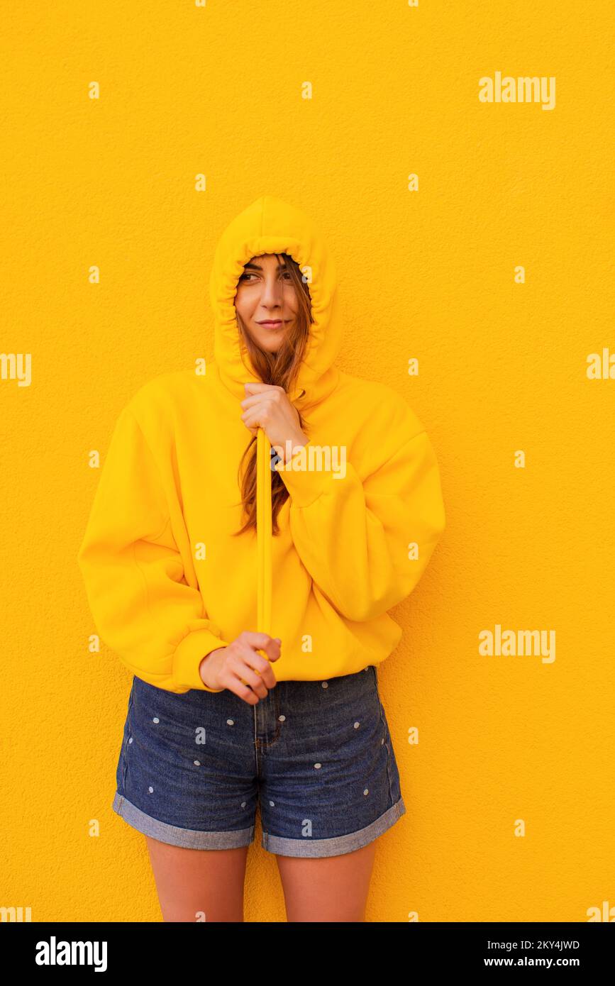 Smiling casual young woman in a yellow hoodie is having fun standing on a colorful yellow background Stock Photo