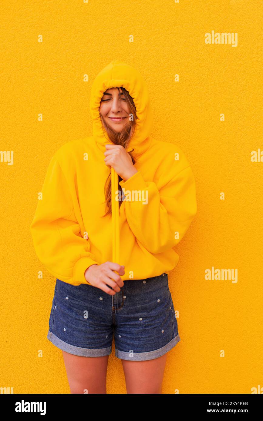 Smiling casual young woman in a yellow hoodie is having fun standing on a colorful yellow background Stock Photo